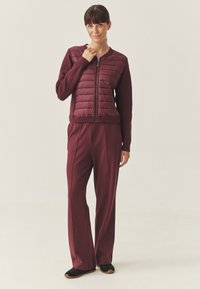 Giacca bordeaux con zip frontale e parte superiore trapuntata, abbinata a pantaloni larghi coordinati. Tessuto morbido con polsini a coste e cuciture laterali. Scarpe nere.