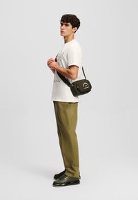 Grøn crossbody taske med lynlås og logo, bæres med en hvid graphic t-shirt og olivengrønne bukser, parret med mørkegrønne sneakers.