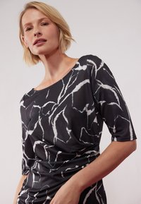 Svart topp med ett marmorerat vitt mönster, tillverkad av ett stretchy material, med båturringning och ärmar som når armbågen.