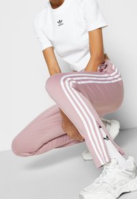 Ljusrosa träningsbyxor med vita ränder längs sidorna, avsmalnande design och dragkedja vid anklarna. Kombinerat med en vit kortärmad tröja och sneakers.