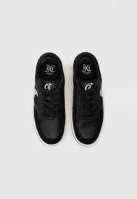 Para czarnych niskich sneakersów z białymi podeszwami, sznurówkami oraz logo "Just Cavalli Milano" wewnątrz strefy pięty na białym tle.