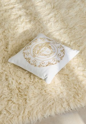 Square white decorative pillow featuring gold intricate embroidery of a face and ornate circular pattern, resting on a textured cream rug.