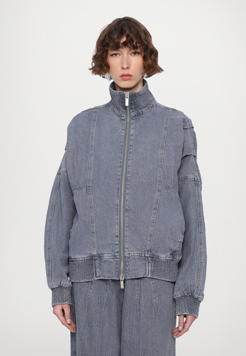 Jachetă din denim gri cu guler înalt, fermoar frontal și manșete cu nervuri. Croială lejeră, detalii matlasate pe umeri și la tiv.
