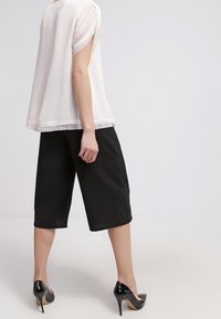 Blusa bianca semi-trasparente con maniche arrotolate, abbinata a culotte nere al ginocchio e tacchi lucidi neri.