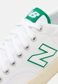 Baskets en toile blanche avec des lacets blancs, une bordure verte, un logo "N" vert et blanc sur le côté et une étiquette verte sur la languette.