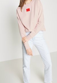 Felpa rosa chiaro con logo rettangolare rosso, abbinata a jeans azzurri ricamati con motivi delicati. Tessuto morbido e vestibilità comoda.