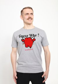 Šedé bavlněné tričko s velkým červeným grafickým motivem krávy a textem „Hádej kdo?“ a „La vache qui rit“ pod tím. Krátké rukávy, volný střih.