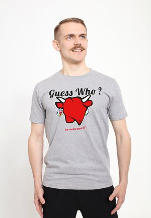Graues Baumwoll-T-Shirt mit einem großen roten Kuh-Graphic und dem Text "Rat mal wer?" sowie "La vache qui rit" darunter. Kurzärmelig, lockere Passform.