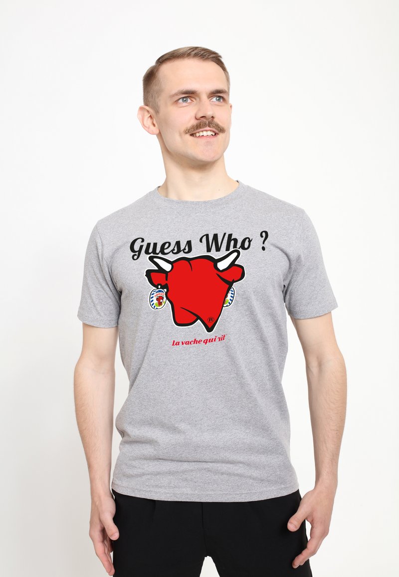 Šedé bavlněné tričko s velkým červeným grafickým motivem krávy a textem „Hádej kdo?“ a „La vache qui rit“ pod tím. Krátké rukávy, volný střih.