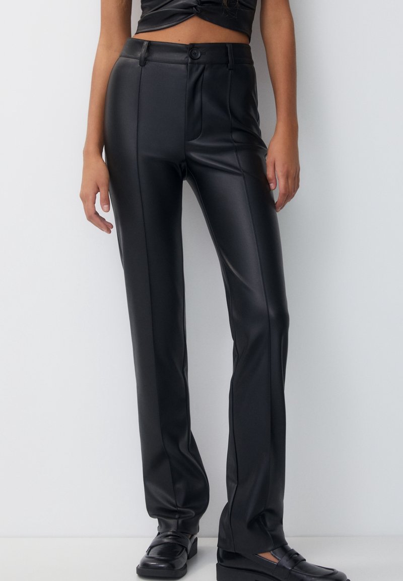 Femme portant un pantalon droit noir en similicuir taille haute associé à des mocassins noirs, debout contre un mur uni.