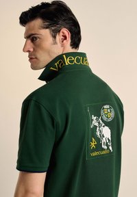Polo verde con el texto amarillo "valecuatro" en el collar, que presenta un gran parche bordado de un jinete y emblema en la parte posterior.