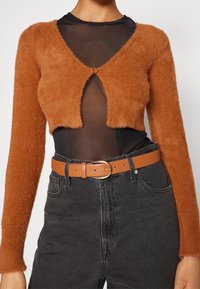 Krzaczasty, przycięty kardigan w kolorze rdzy na przejrzystej czarnej bluzce, zestawiony z czarnymi jeansami z wysokim stanem i jasnobrązowym paskiem ze złotą klamrą.