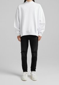 Vit långärmad sweatshirts med hög krage, avslappnad passform och ribbade ärmslut, kombinerad med rivna svarta skinny jeans och vita sneakers.