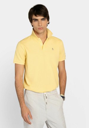 Giovane uomo con una polo gialla a maniche corte e pantaloni chiari a righe con coulisse, in piedi davanti a uno sfondo chiaro e uniforme.