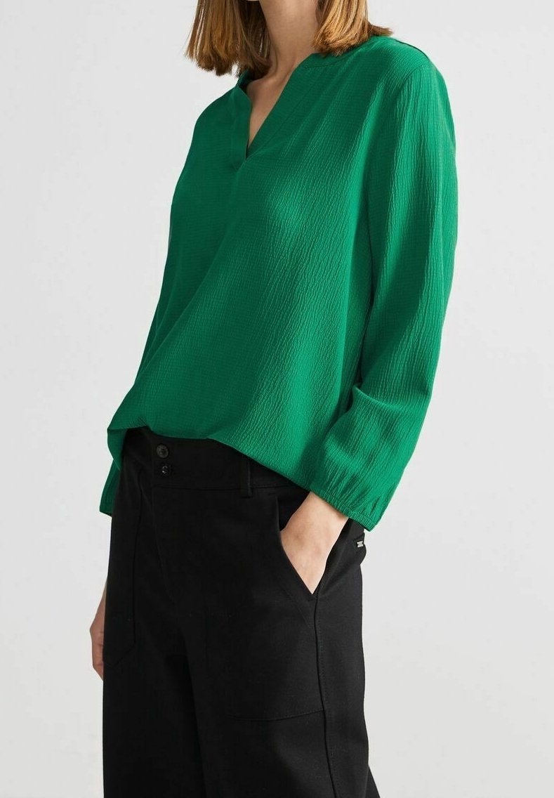 Femme portant un chemisier vert texturé à manches longues avec un col en V et un pantalon noir, une main dans la poche, sur un fond uni.