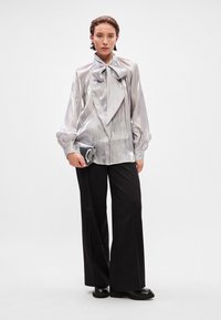 Silver, shiny blouse with a large bow at the collar, long sleeves, and textured fabric. Paired with black, wide-legged trousers and a clutch.