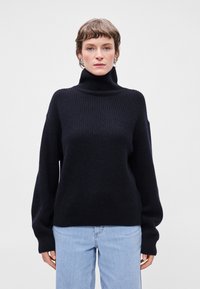 Σκούρο ναυτικό oversized πουλόβερ με ψηλό γιακά, ριγωτή υφή και μακριά, φαρδιά μανίκια, συνδυασμένο με ανοιχτό καφέ τζιν.