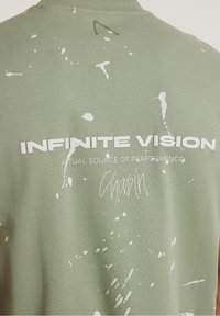 Olive green t-shirt featuring white splatter patterns and bold text: "INFINITE VISION" with "ACTUAL SOURCE OF PERFORMANCE" below.