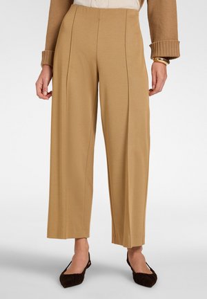 Pantaloni beige a gamba larga indossati con un maglione marrone, un braccialetto d'oro e ballerine nere con punta su una persona in piedi davanti a uno sfondo bianco.