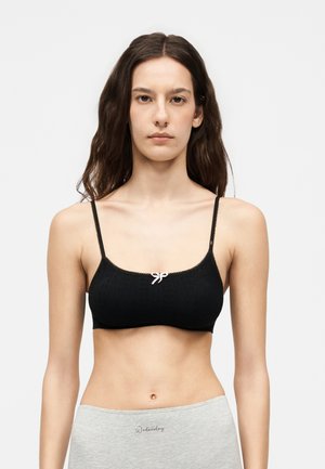 Νέα γυναίκα με μακριά σκούρα μαλλιά που φοράει ένα μαύρο κοντό τοπ με λεπτές τιράντες και ένα μικρό φιόγκο, καθώς και ανοιχτό γκρι παντελόνι με το κείμενο "Wednesday".