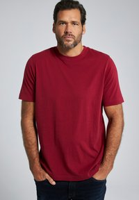 Rode katoenen t-shirt met ronde hals en korte mouwen. De stof heeft een gladde textuur. Gecombineerd met donkere spijkerbroeken, handen in de zakken. Neutrale achtergrond.