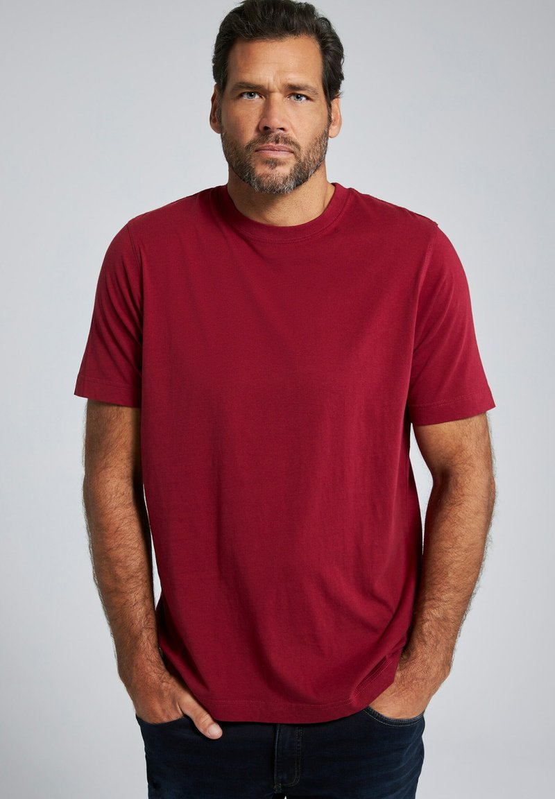 Rode katoenen t-shirt met ronde hals en korte mouwen. De stof heeft een gladde textuur. Gecombineerd met donkere spijkerbroeken, handen in de zakken. Neutrale achtergrond.
