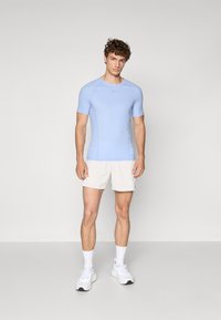 Helles blaues Kurzarm-Sportshirt mit taillierter Passform und strukturierten Seitenpanelen, kombiniert mit beigen Shorts und weißen Sportschuhen.