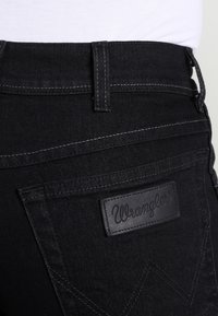 Black denim jeans with a textured finish, featuring a leather logo patch, contrast stitching, and distinctive pocket design.