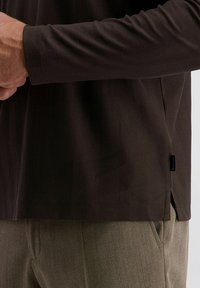 Langarmhemd mit braunem Fischgrätenmuster, locker geschnitten und mit Seitenschlitzen, kombiniert mit strukturierten, hellgrünen Hosen.