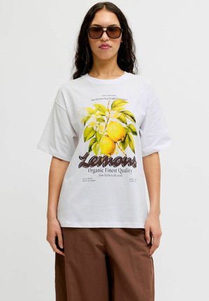 Žena so zatmavenými slnečnými okuliarmi, v bielom tričku s dizajnom žltých citrónov a zelených listov a vhnedých nohaviciach na jednoduchom pozadí.