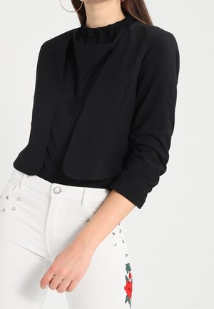 Femme portant une veste courte noire à manches longues et un pantalon blanc avec une broderie florale rouge sur la cuisse.