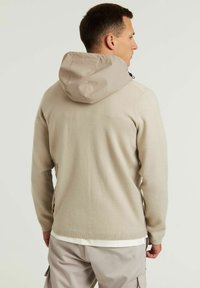 Sudadera con capucha, material de punto beige, acabado texturizado, bolsillo tipo canguro y cordón ajustable en la capucha, diseño sin costuras en los hombros.