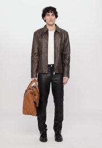 Bruine leren jas, witte shirt, zwarte leren broek en zwarte schoenen. Houdt een tan tas met verstelbare banden vast. Eenvoudig ontwerp.