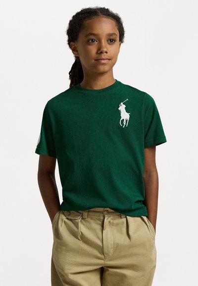 Grünes Baumwoll-T-Shirt mit kurzen Ärmeln, mit einem weißen Polospieler-Logo auf der linken Brust. Getragen mit khakifarbenen Hosen.