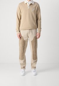 Beige långärmad polo med krage och logotyp; tillsammans med beige byxor som har bruna kordflöjlsfickor fram. Vita sneakers.
