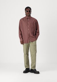 Tjekket bordeaux-knapshirt med en lomme, parret med lysegrønne bukser og sorte kraftige sko. Modellen har runde solbriller på.
