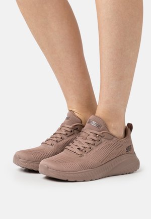 Baskets de sport marron clair à lacets avec dessus en maille texturée portées pieds nus sur un fond blanc uni.