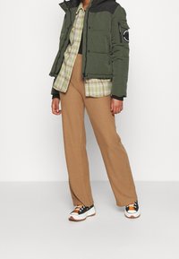 Doudoune verte avec accents noirs, chemise à carreaux vert et beige, pantalon beige à larges jambes côtelées, et baskets noir et blanc avec orange.