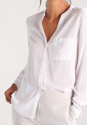 Witte blouse met lange mouwen en knopen, met borstzak, gedragen in lichtgekleurde broek, hand in broekzak tegen een neutrale achtergrond.