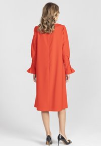 Czerwona sukienka z długim rękawem zakończona rozkloszowanymi mankietami, o gładkiej fakturze, sięgająca do kolan, z centralnym szwem z tyłu. Noszona z czarnymi szpilkami.