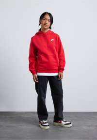 Hanorac roșu cu glugă, cu un buzunar frontal și un logo alb, asortat cu blugi negri din denim și adidași multicolori, pe o suprafață gri.