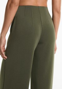 Olive zelené nohavice s širokými nohavicami z mäkkého, elastického materiálu. Majú vysoký pás s jemnými plisovaním a bočnými vreckami. Hladká textura po celom povrchu.