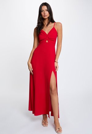 Donna in piedi che indossa un abito rosso senza maniche con apertura frontale, spacco alto sulla coscia e sandali con cinturini su uno sfondo chiaro e semplice.
