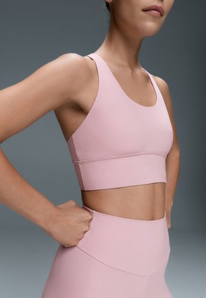Femme en brassière de sport rose et leggings avec les mains sur les hanches, montrée des épaules jusqu’aux hanches sur un fond gris uni.
