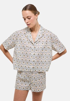 Femme avec les cheveux en chignon portant une chemise à manches courtes et un short assortis à motif floral sur un fond clair, les mains posées sur les hanches.