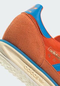 Oranje suede sneaker met blauwe accenten, voorzien van een gestructureerde bovenkant, gouden branding en een crèmekleurige rubberen zool met profiel.