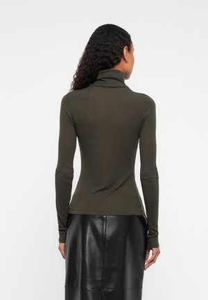 Frau mit lockigem Haar trägt einen olivgrünen Rollkragenpullover und einen schwarzen Lederrock bis zum Knie, sie steht mit dem Rücken zu einem weißen Hintergrund.