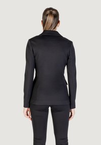 Donna con una coda di cavallo biondo scuro, che indossa un blazer nero aderente e pantaloni abbinati, in piedi contro uno sfondo chiaro e uniforme.