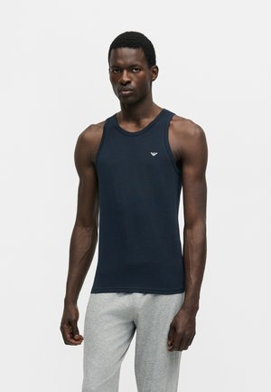 Man wearing a fitted navy sleeveless tank top with a small logo on chest and light gray sweatpants, standing against a plain background.