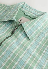 Chemise à carreaux verts et bleu clair avec col et fermeture éclair métallique à l'avant, affichant une étiquette "NEXT EDIT." à l'intérieur du col.